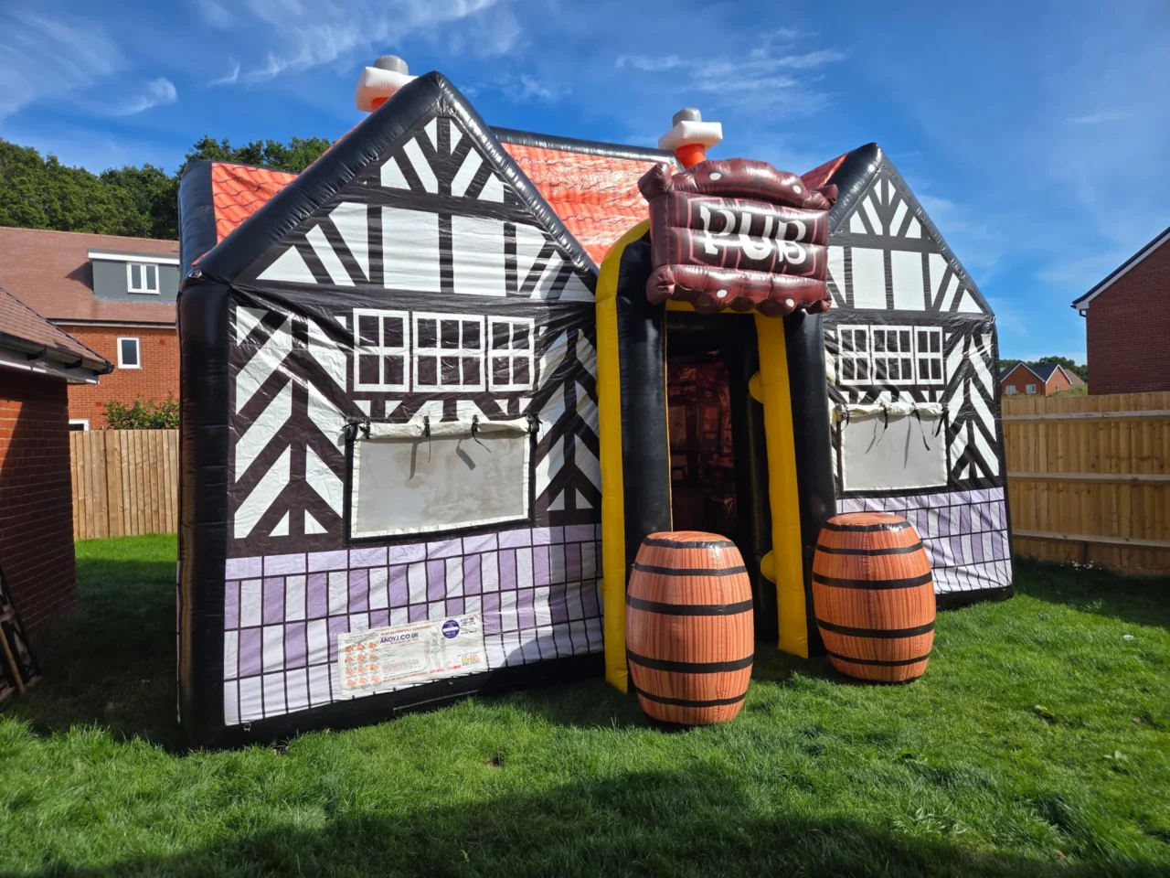 Black-and-white inflatable pub with an orange roof and barrel props, set up on grass in a garden